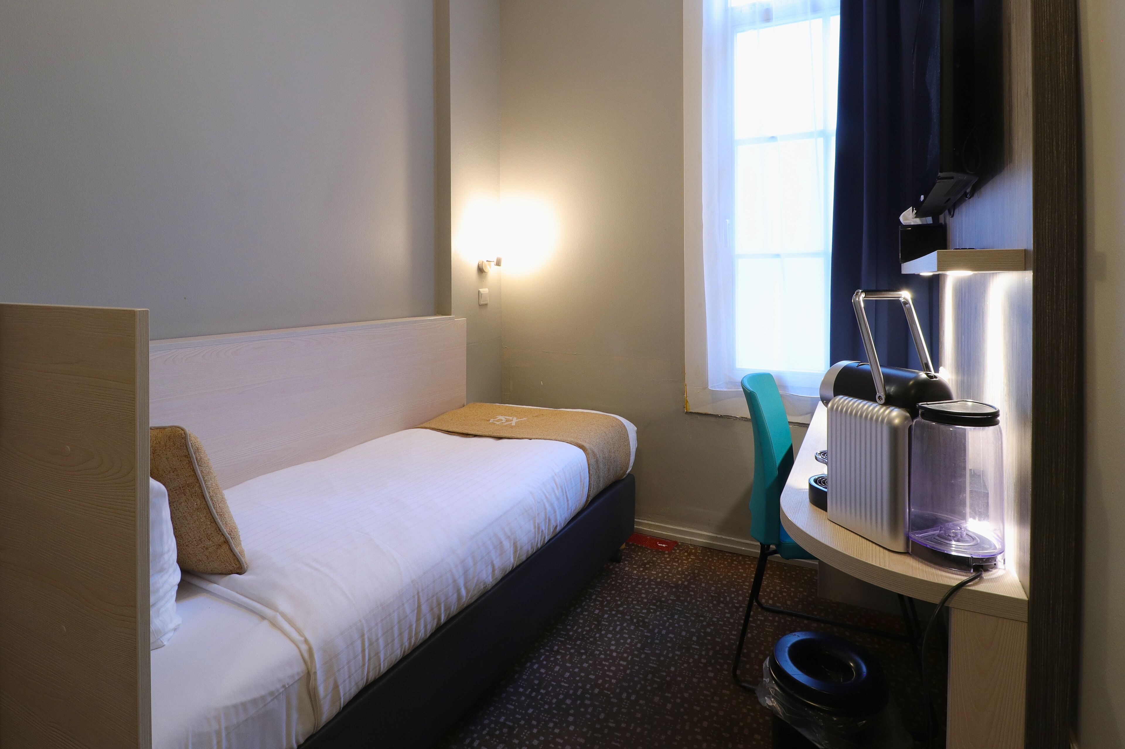 A hotel room with a bed, a nightstand with a lamp, a window with curtains, and a wall-mounted TV.