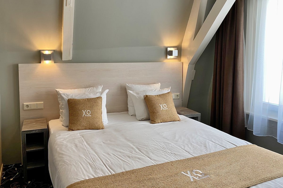 Een hotelkamer met een groot bed, twee nachtkastjes, een hoofdbord met ingebouwde verlichting en uitzicht op het landschap door een raam met dunne gordijnen.