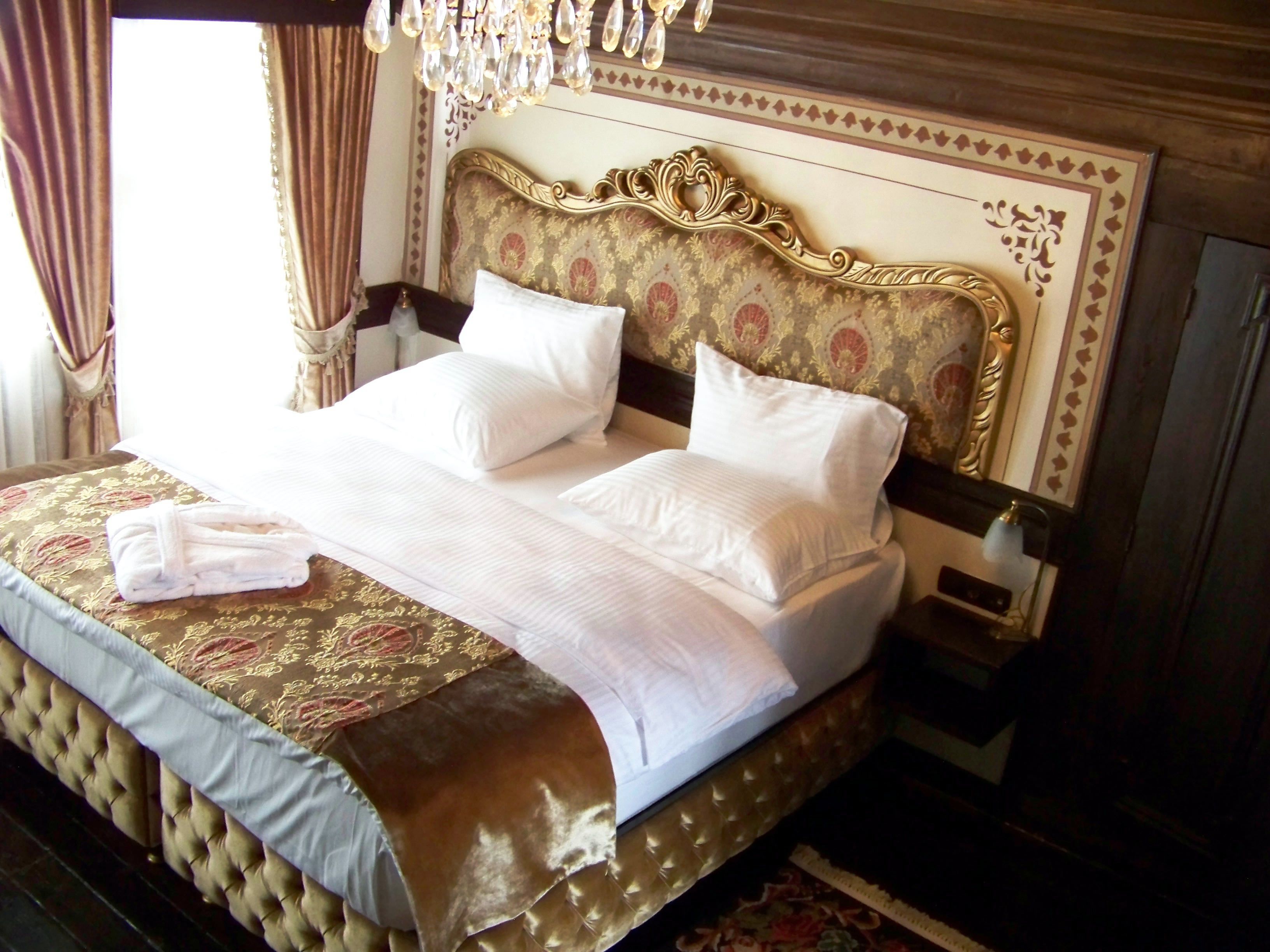 Un grand lit avec une tête de lit dorée, du linge de lit blanc et une robe pliée posée sur une table de chevet.