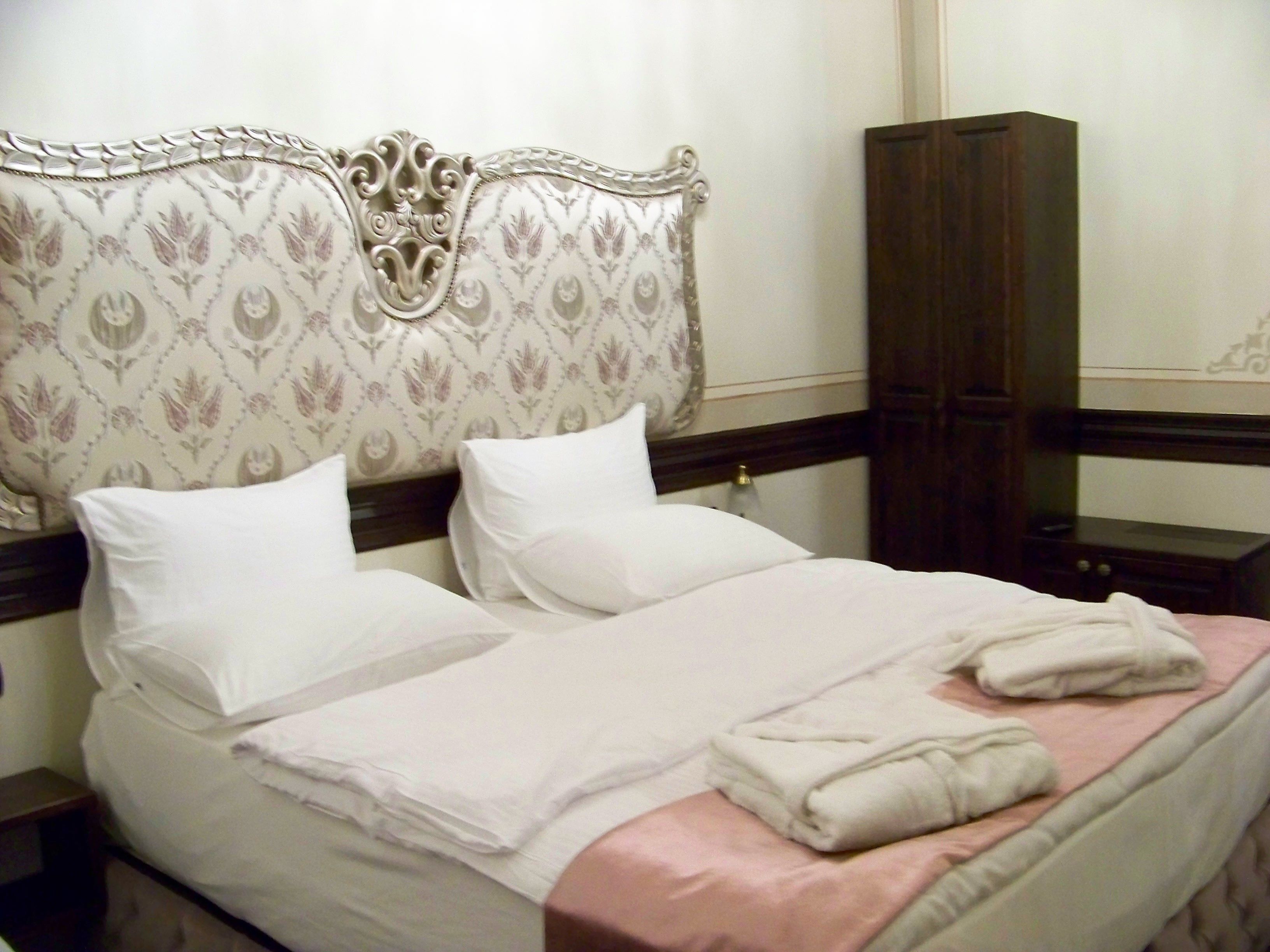 Una cama bien tendida con sábanas blancas y un toque de color rosa, una mesita de noche de madera y un armario de madera oscura.