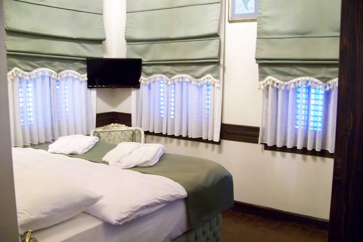 Una cama bien hecha con almohadas y una toalla doblada, un televisor montado en la pared y una ventana con cortinas.