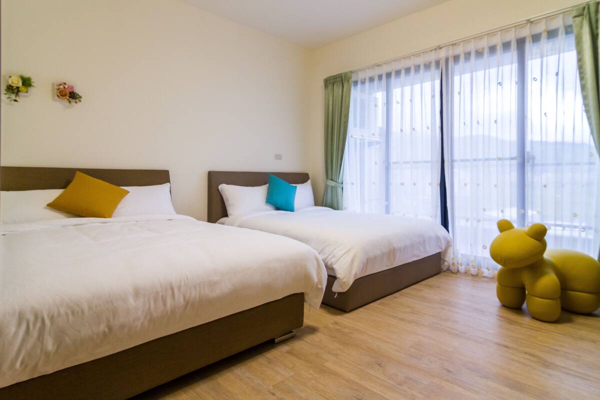 Une chambre d’hôtel avec deux lits, une grande fenêtre avec des rideaux légers et un coussin en forme d’ours posé sur le sol.