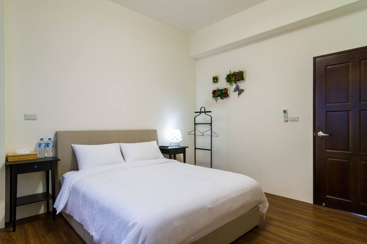 Un lit bien fait, avec du linge de lit blanc, une table de chevet avec une lampe et un plancher en bois.