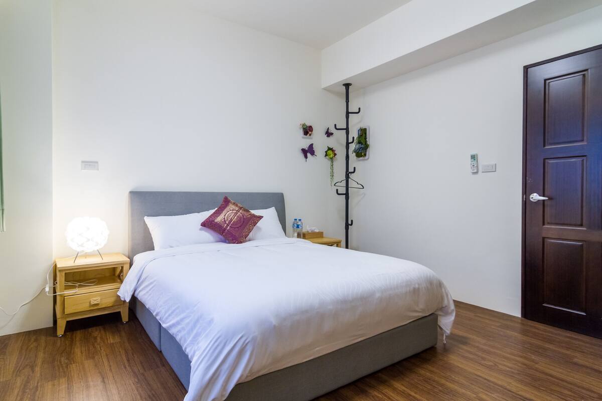 Un lit bien fait, avec du linge de lit blanc, une table de chevet en bois surmontée d’une lampe et une plante fixée au mur.