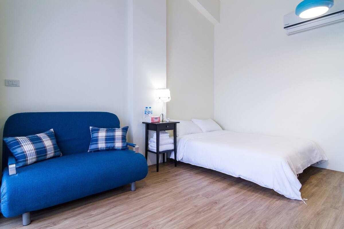 Une pièce avec un canapé bleu, un lit blanc, une petite table avec une lampe et un climatiseur fixé au mur.