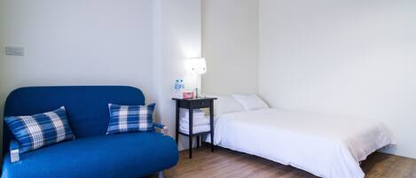Une pièce avec un canapé bleu, un lit blanc, une petite table avec une lampe et un climatiseur fixé au mur.