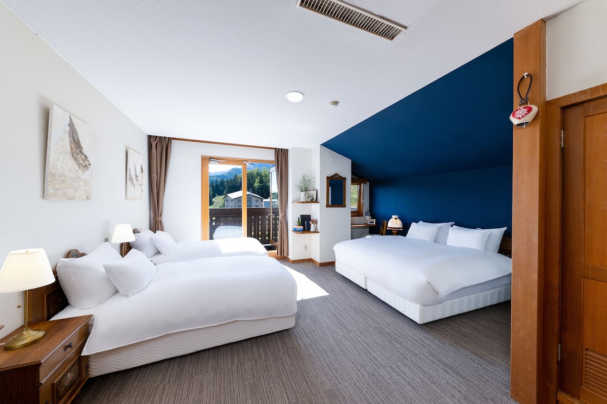 Habitación de hotel con dos camas, una mesita de noche de madera, una lámpara, una ventana con vistas a montañas y una pared azul decorativa.