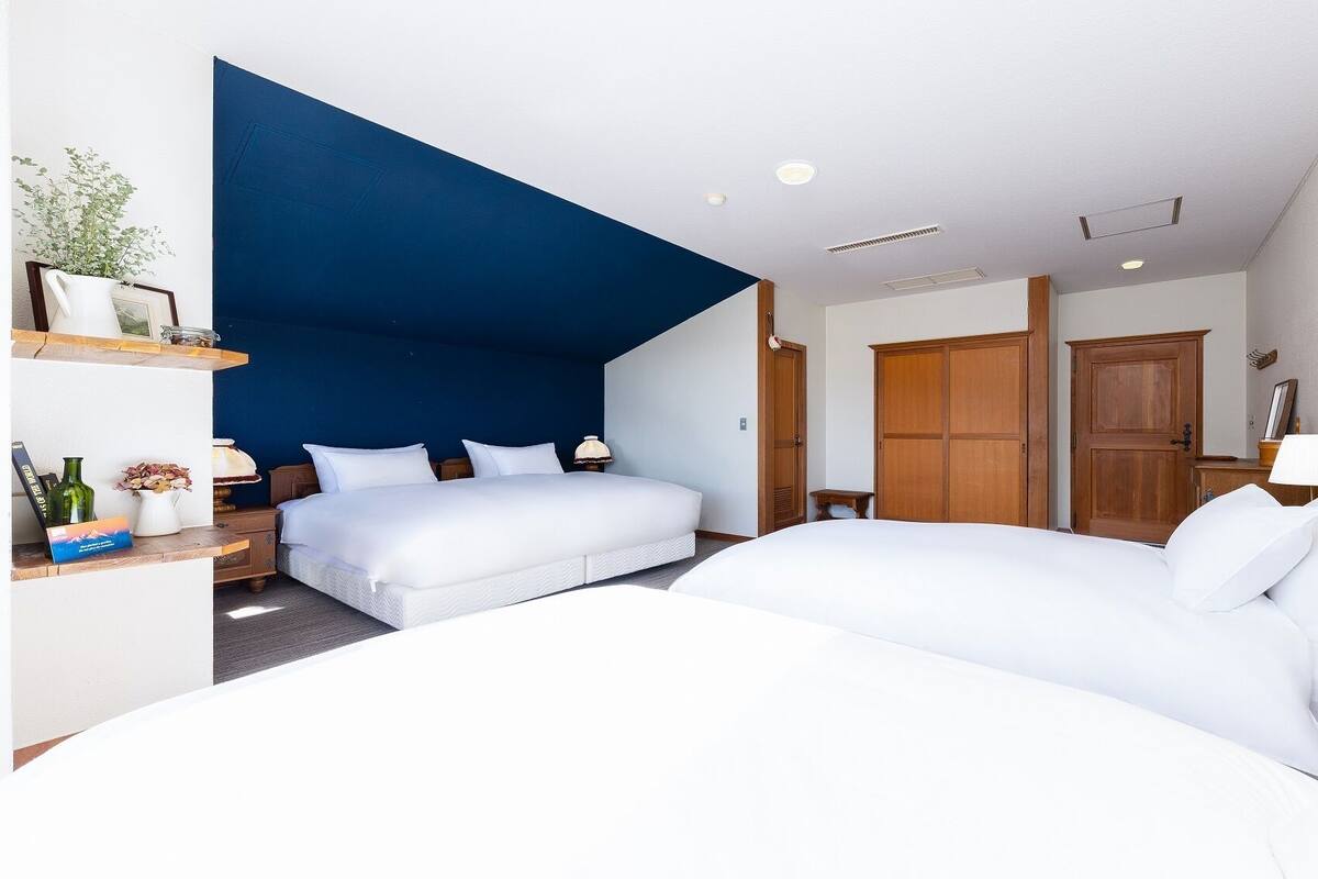 Habitación de hotel con dos camas, una pared de acento azul, armario de madera y una pequeña mesa con lámpara.