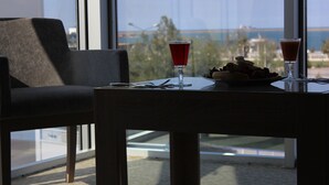 Uma mesa com duas bebidas e um prato de comida, uma cadeira e uma janela grande com vista para o mar e edifícios.