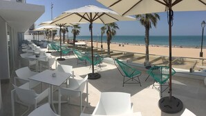 Una terraza con mesas y sillas blancas, sombrillas grandes y vistas a la playa y al océano.