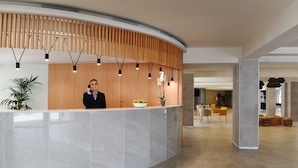 Un mostrador de recepción de diseño moderno, con encimera de mármol y listones de madera en la parte superior. Hay una planta en maceta y un jarrón con flores sobre el mostrador. La zona de recepción da paso a un comedor con sillas amarillas y una mesa.