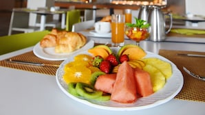 Una escena de desayuno con un plato de fruta y cruasanes, una cafetera y un vaso de jugo en una mesa, en un comedor moderno con vigas de madera en el techo.