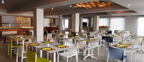 A modern dining area with tables and chairs, a kitchen in the background, and a ceiling with wooden geometric designs.