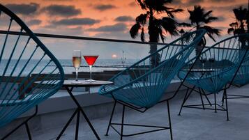 Eine Terrasse mit blauen Drahtstühlen und einem Tisch, mit Blick auf das Meer, Palmen und einen Sonnenuntergang.