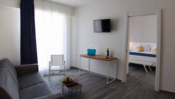 A modern hotel room with a grey sofa, a small table with a basket, a TV mounted on the wall, and a bed with yellow pillows.
