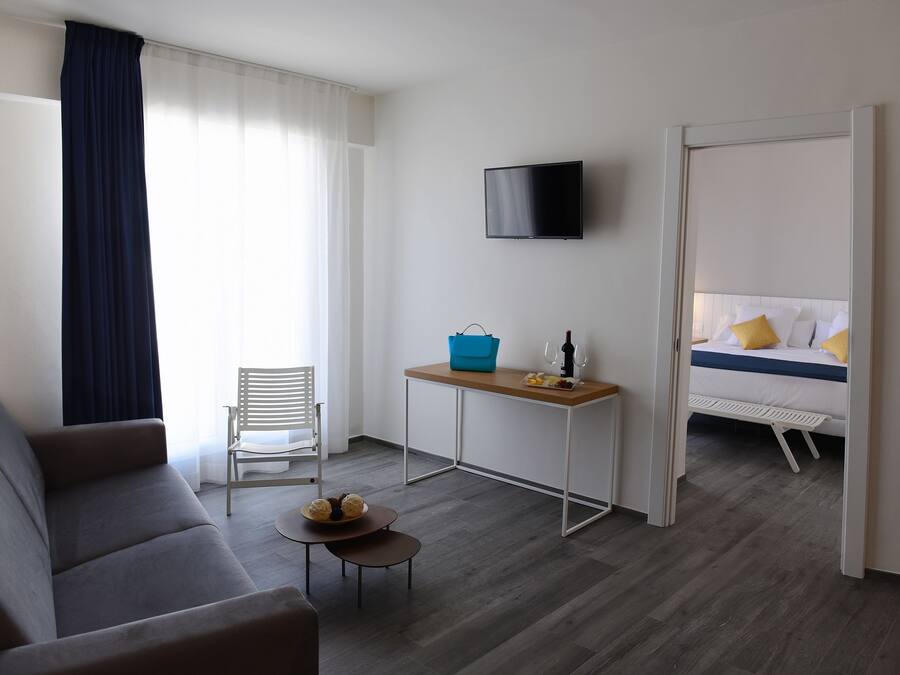 A modern hotel room with a grey sofa, a small table with a basket, a TV mounted on the wall, and a bed with yellow pillows.