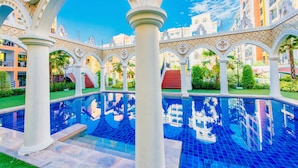 An indoor pool area with white columns and arches, surrounded by greenery and buildings in the background.