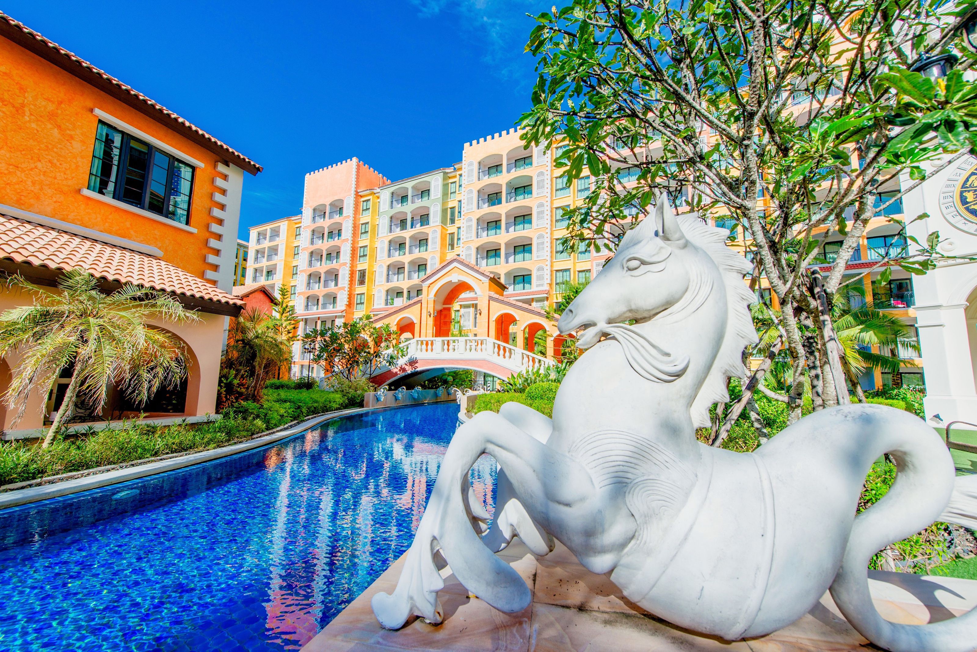 A swimming pool with a white horse statue, surrounded by buildings with balconies and a bridge.