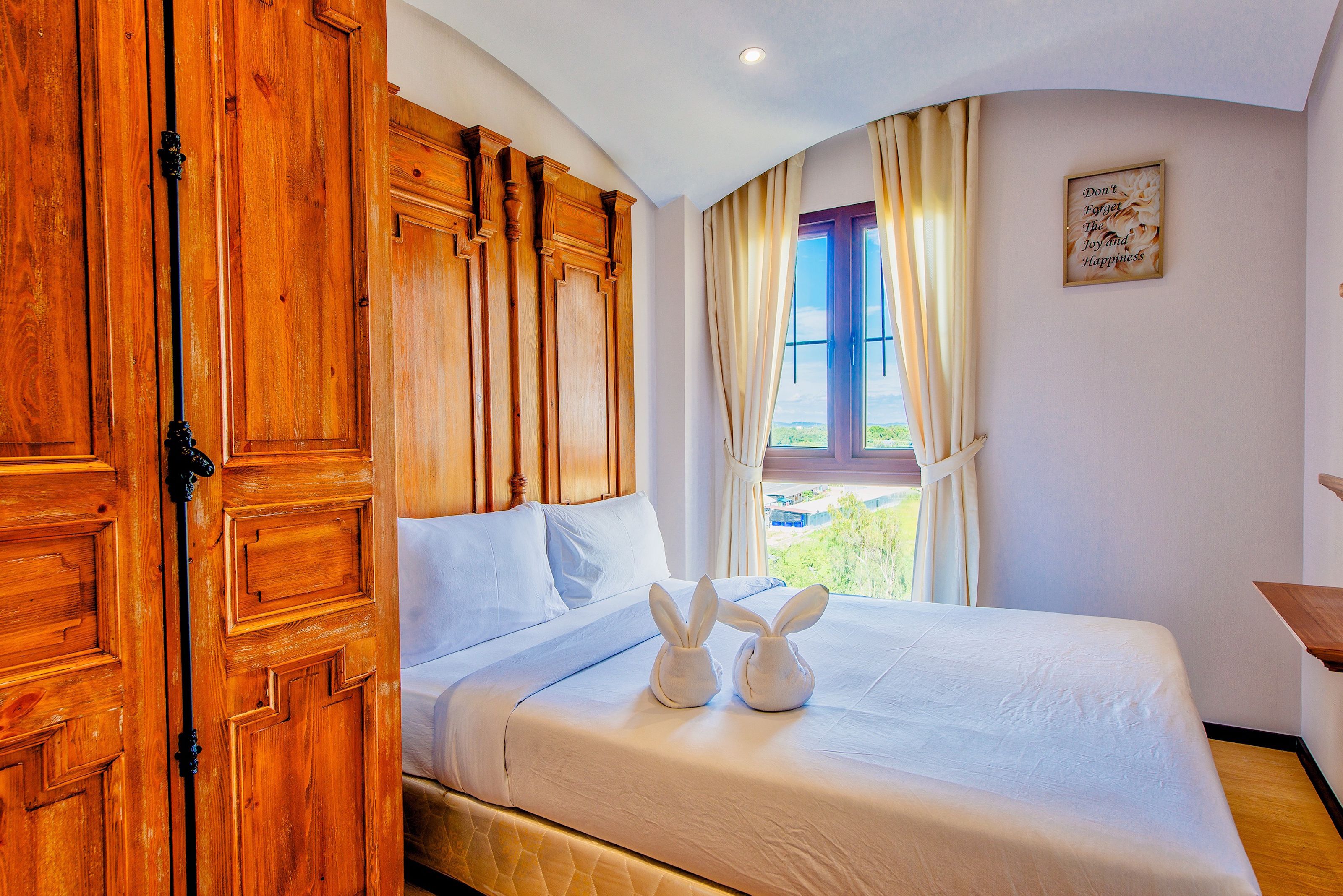 A neatly made bed with white bedding and two white bunny-shaped pillows, a wooden wardrobe, and a window with curtains.