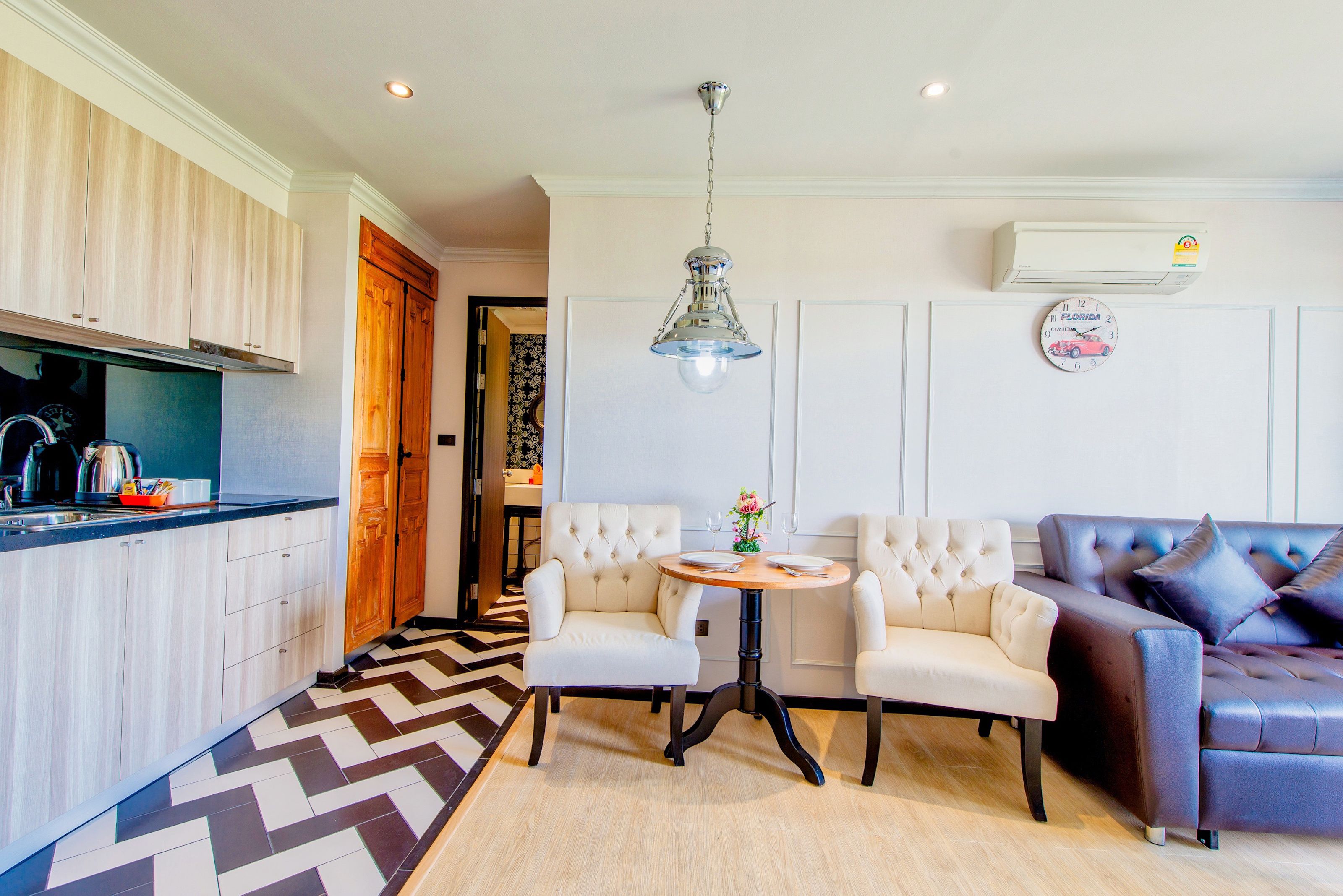 Una cocina moderna con armarios blancos, un suelo con estampado blanco y negro, una zona de comedor con dos sillas blancas y una mesa redonda, y un sofá de cuero marrón oscuro.