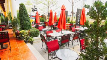An outdoor cafe with red chairs and tables, surrounded by potted plants and orange umbrellas.