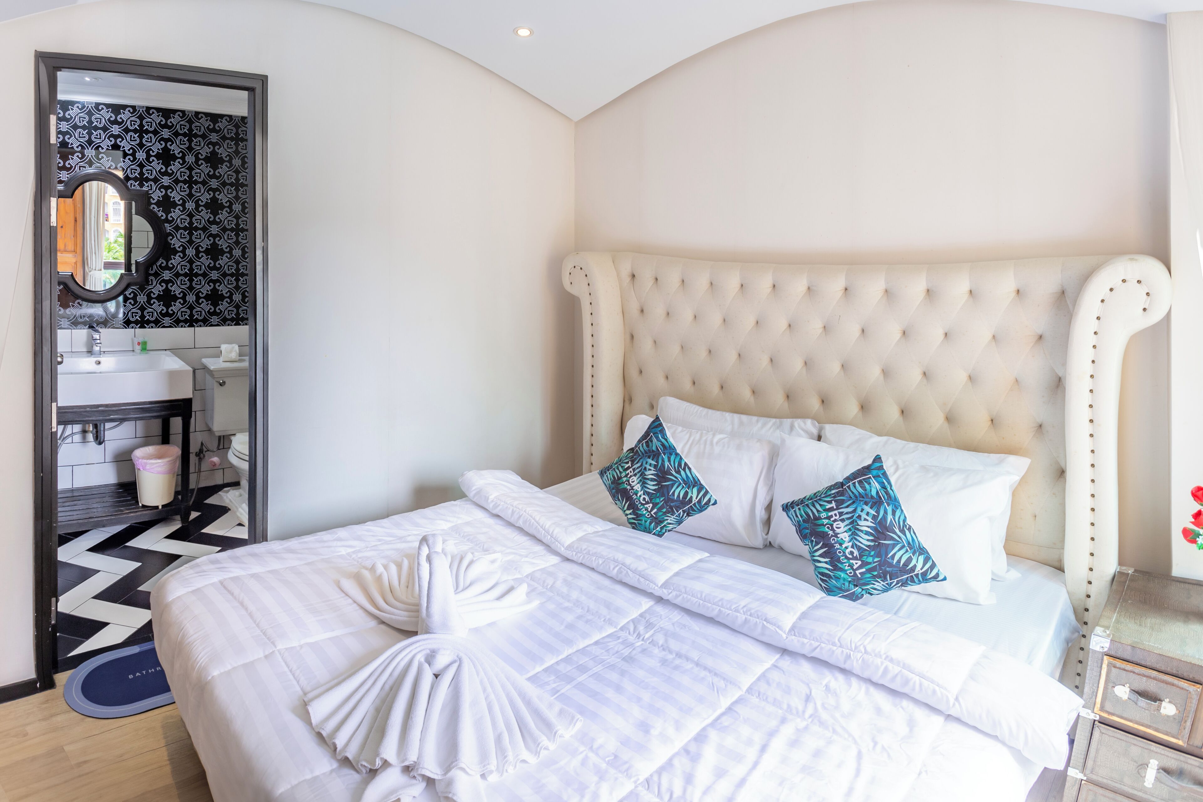 Una cama bien hecha con sábanas blancas y dos almohadones decorativos, un cabecero capitonado y una mesita de noche con una lámpara.