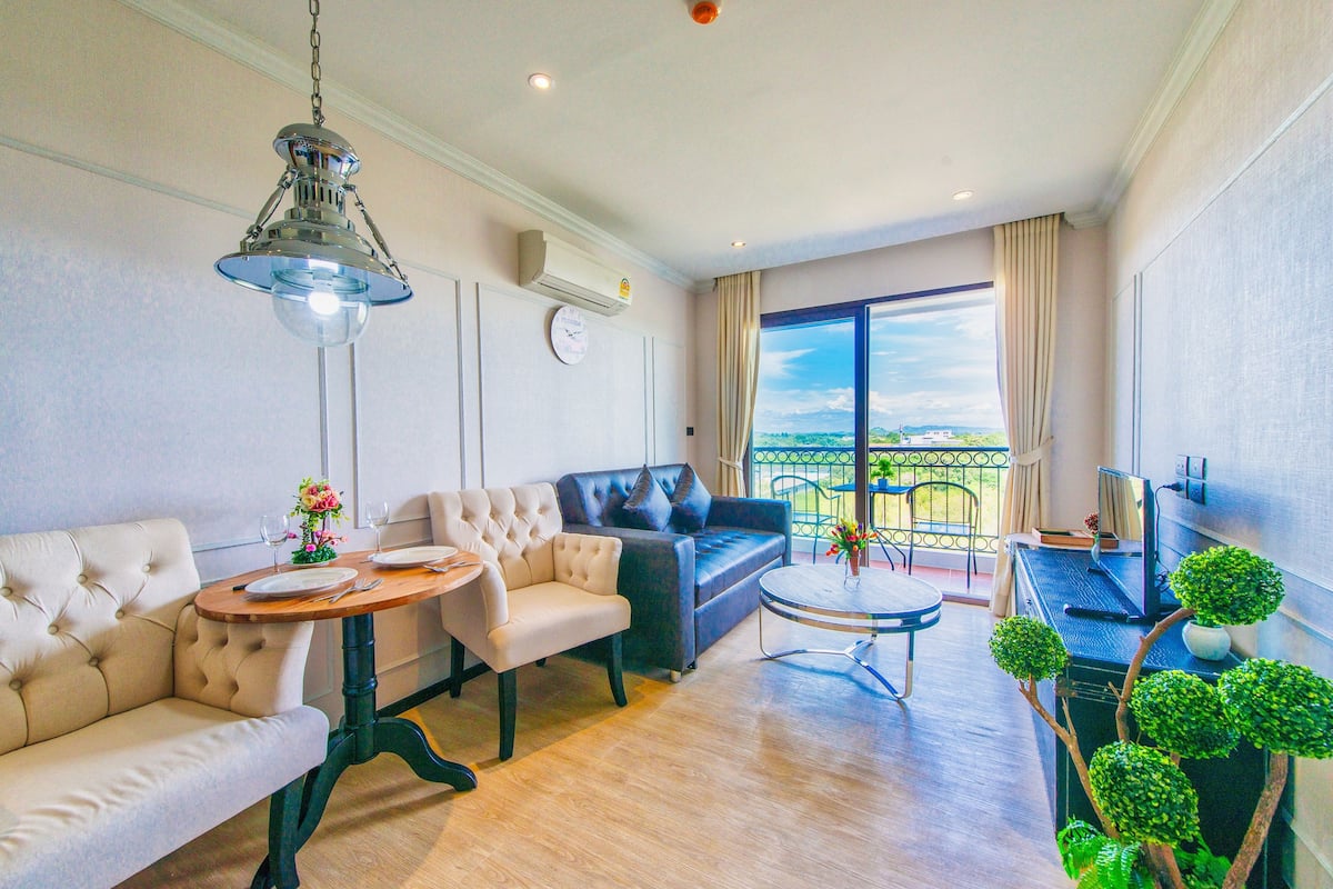Una sala moderna con sofá, sillón y una mesa redonda. Cuenta con un balcón con vistas, un televisor y una pequeña planta.