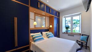 A bedroom with a large bed, two bedside lamps, a window with a view of buildings, and a chair with a cushion. - Maison Lauren (Sorrento)