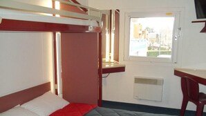 A single bed with a red and gray quilt, a wooden headboard, a small desk with a chair, a window with a view of buildings, and a wall-mounted heater. - Formul'Eco Maubeuge (Maubeuge)