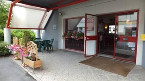 A building entrance with a covered outdoor seating area, a wooden bench with flowers, and a glass door with a red frame. - Formul'Eco Maubeuge (Maubeuge)