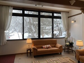 A cozy living room with a large window, a brown leather sofa, two small tables with lamps, and a patterned rug. - Wadano Forest Hotel And Apartments (Hakuba)