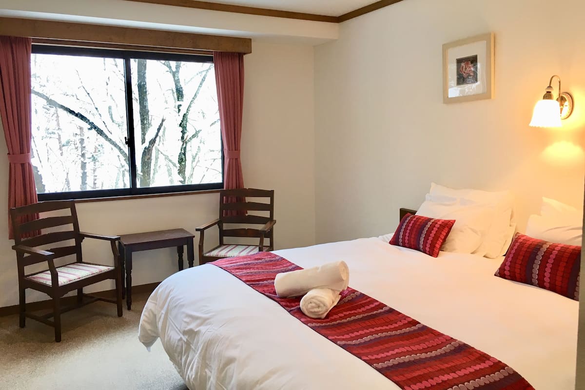 A hotel room with a large bed, a desk, two chairs, a window with curtains, and a wall-mounted light.