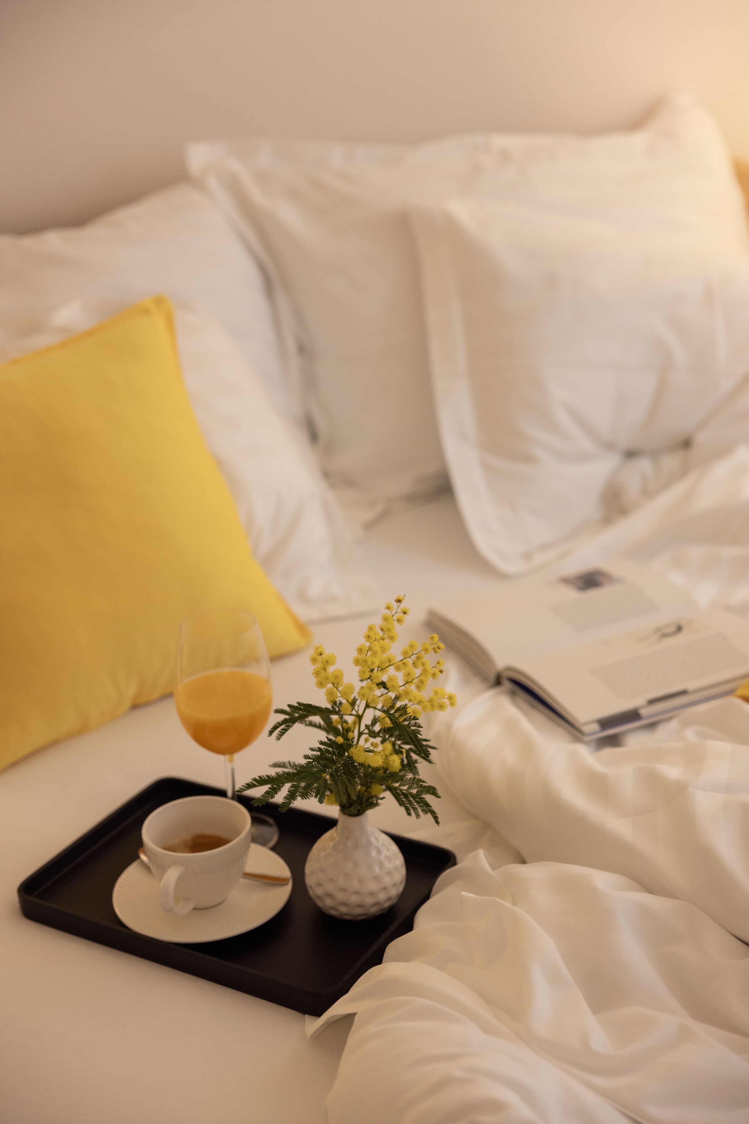 A neatly made bed with white and yellow pillows, a tray with a cup of coffee, a glass of orange juice, and a small vase with yellow flowers.