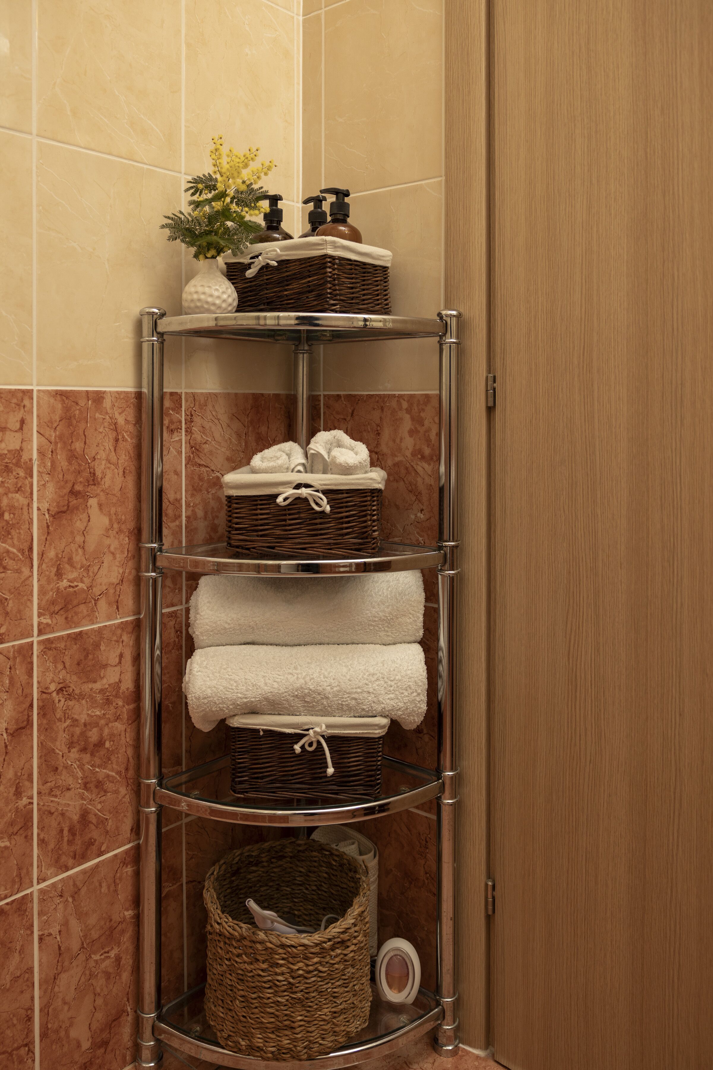 A corner bathroom with a chrome towel rack holding white towels, a wicker basket, and toiletries.