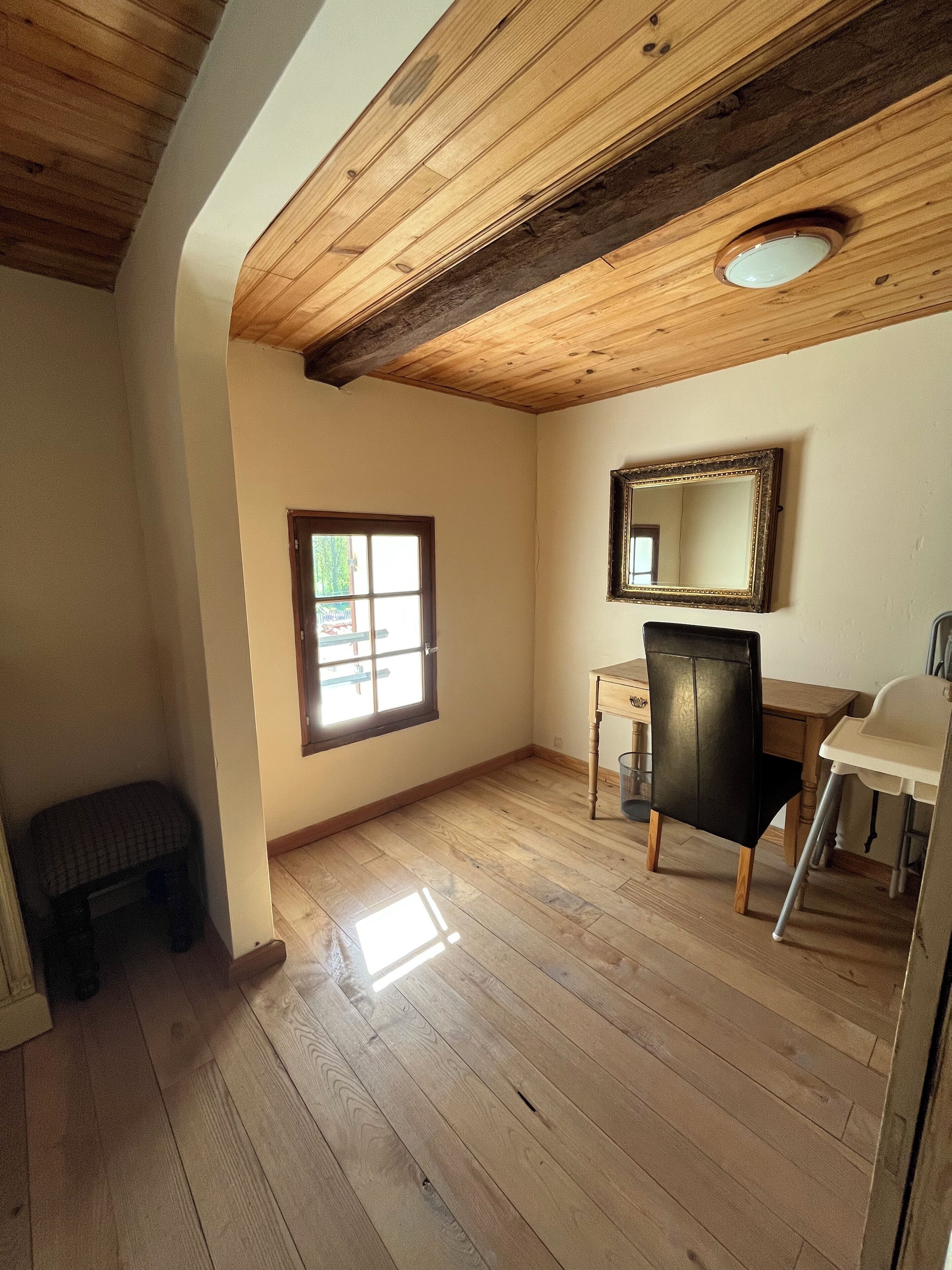 A room with wooden flooring, a window, a desk with a chair, and a mirror on the wall.