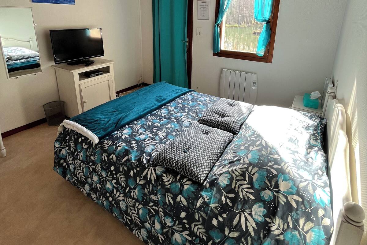 A bedroom with a wooden ceiling, a bed with a floral bedspread, a television on a stand, and a window with curtains.