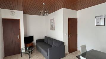 A living room with a wooden ceiling, a gray sofa, a glass coffee table, a TV, a dining table with chairs, and a chandelier.
