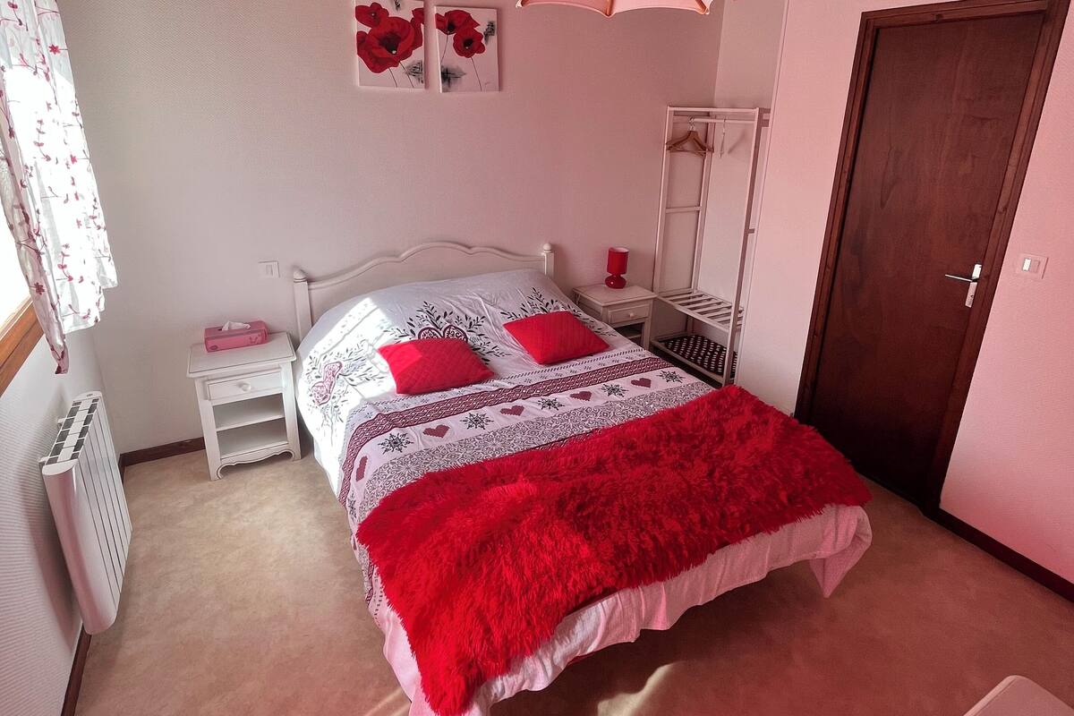 A bedroom with a wooden ceiling, a bed with a red blanket, a nightstand, a radiator, and a door.