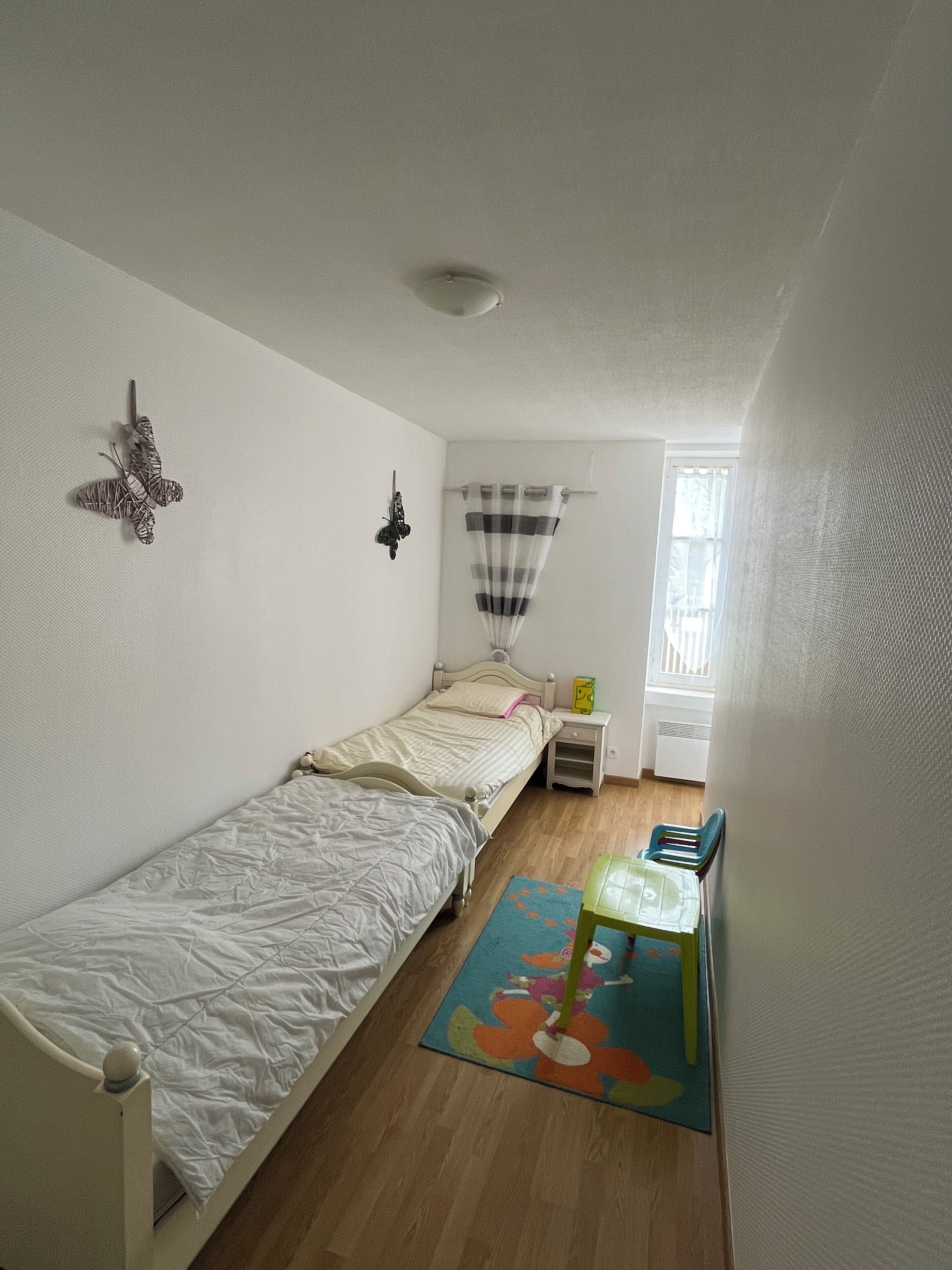 Una piccola camera con un letto singolo, pavimento in legno, una finestra con tenda a righe, un tavolino con una sedia e un tappeto colorato.