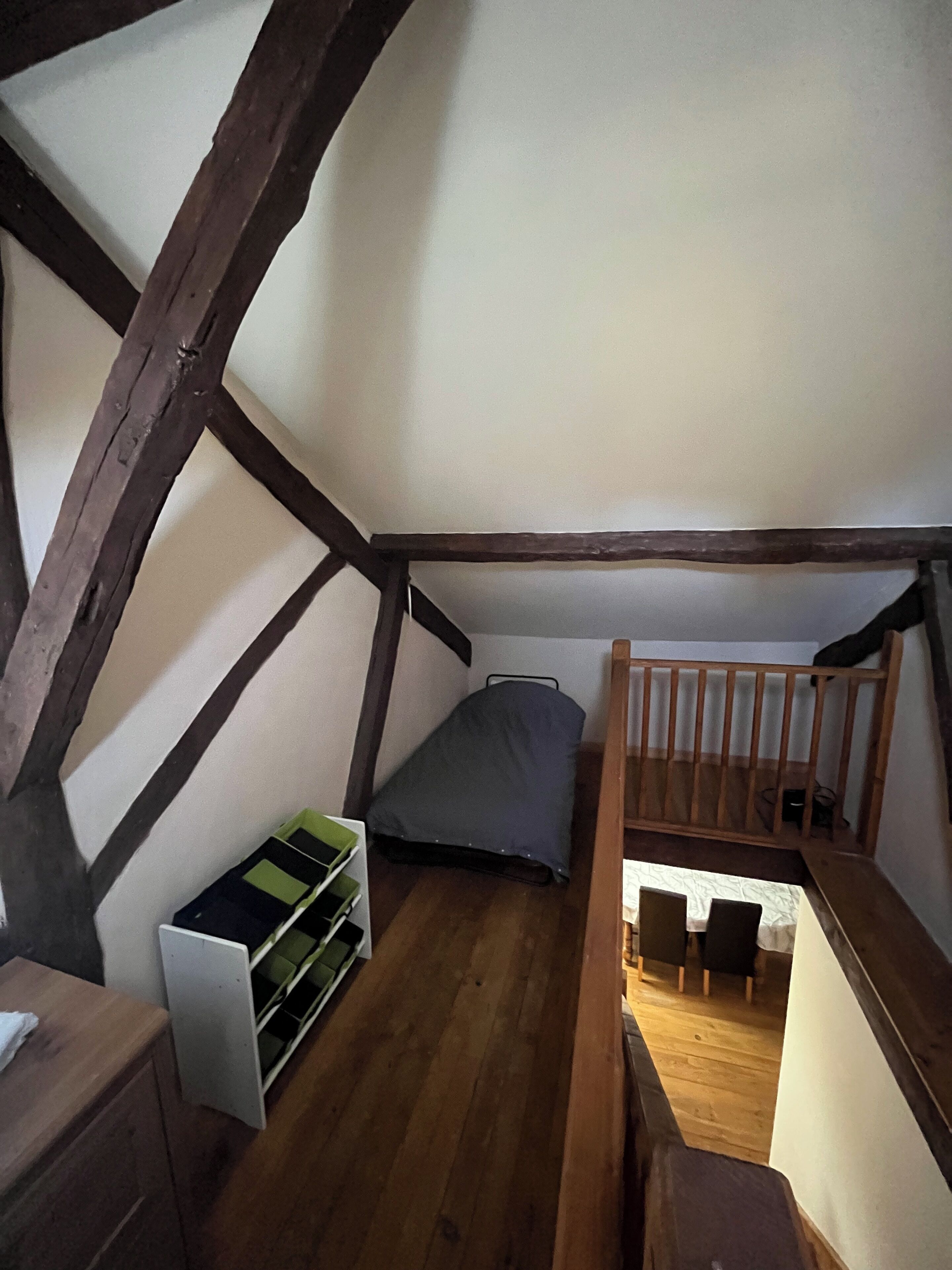 A small attic bedroom with a sloped ceiling, wooden beams, a bunk bed, a bedside table, and a green storage unit.