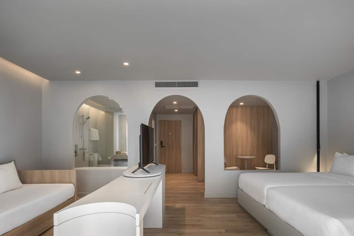 A modern hotel room with a sofa, a bed, a TV, and a bathroom visible through an archway.