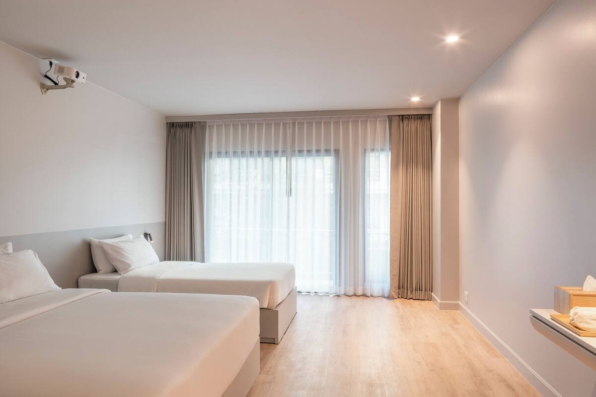 A hotel room with two beds, a large window with curtains, a ceiling-mounted projector, and a bedside table with a lamp.