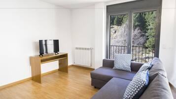 Uma sala de estar moderna com um sofá cinza, uma mesa lateral de madeira, uma televisão e uma janela grande com vista para árvores.