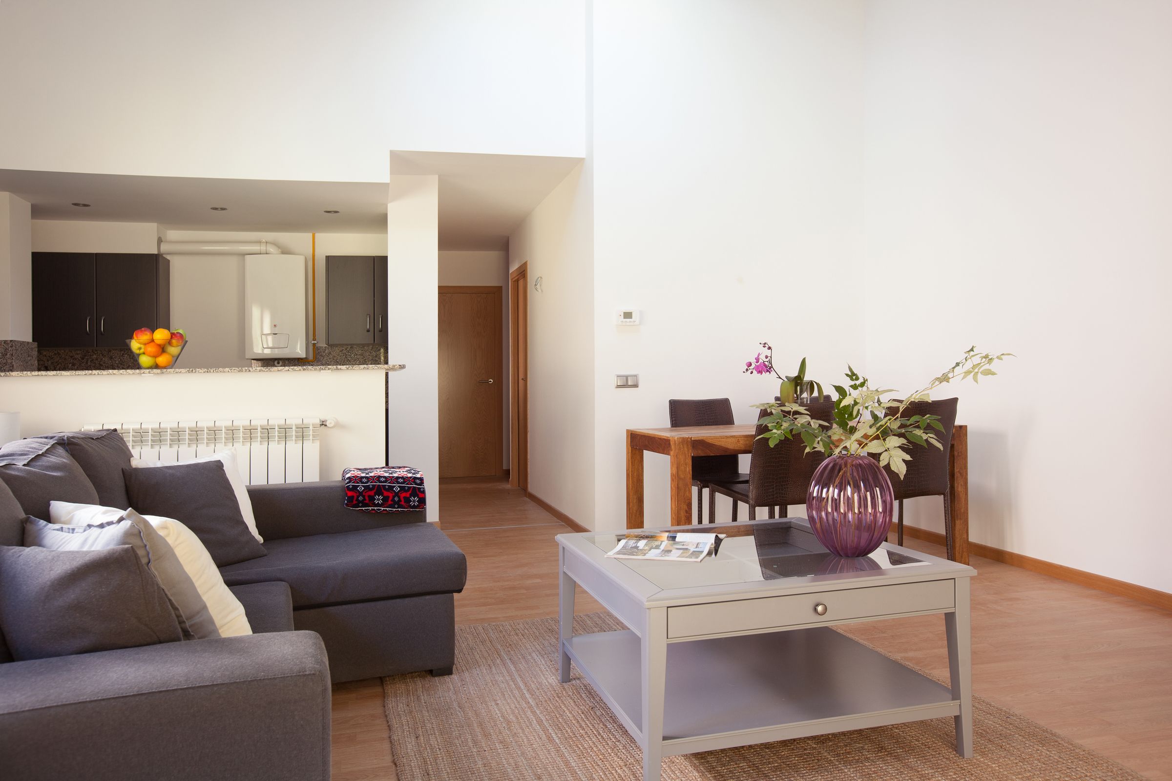 A modern living room with a grey sofa, a small coffee table, and a dining area with a wooden table and chairs.