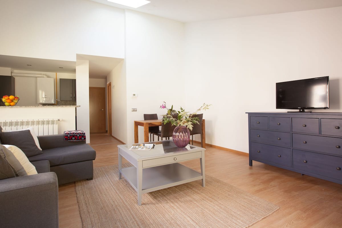 A modern living room with a grey sofa, a coffee table, a TV, and a dining area with chairs and a table.
