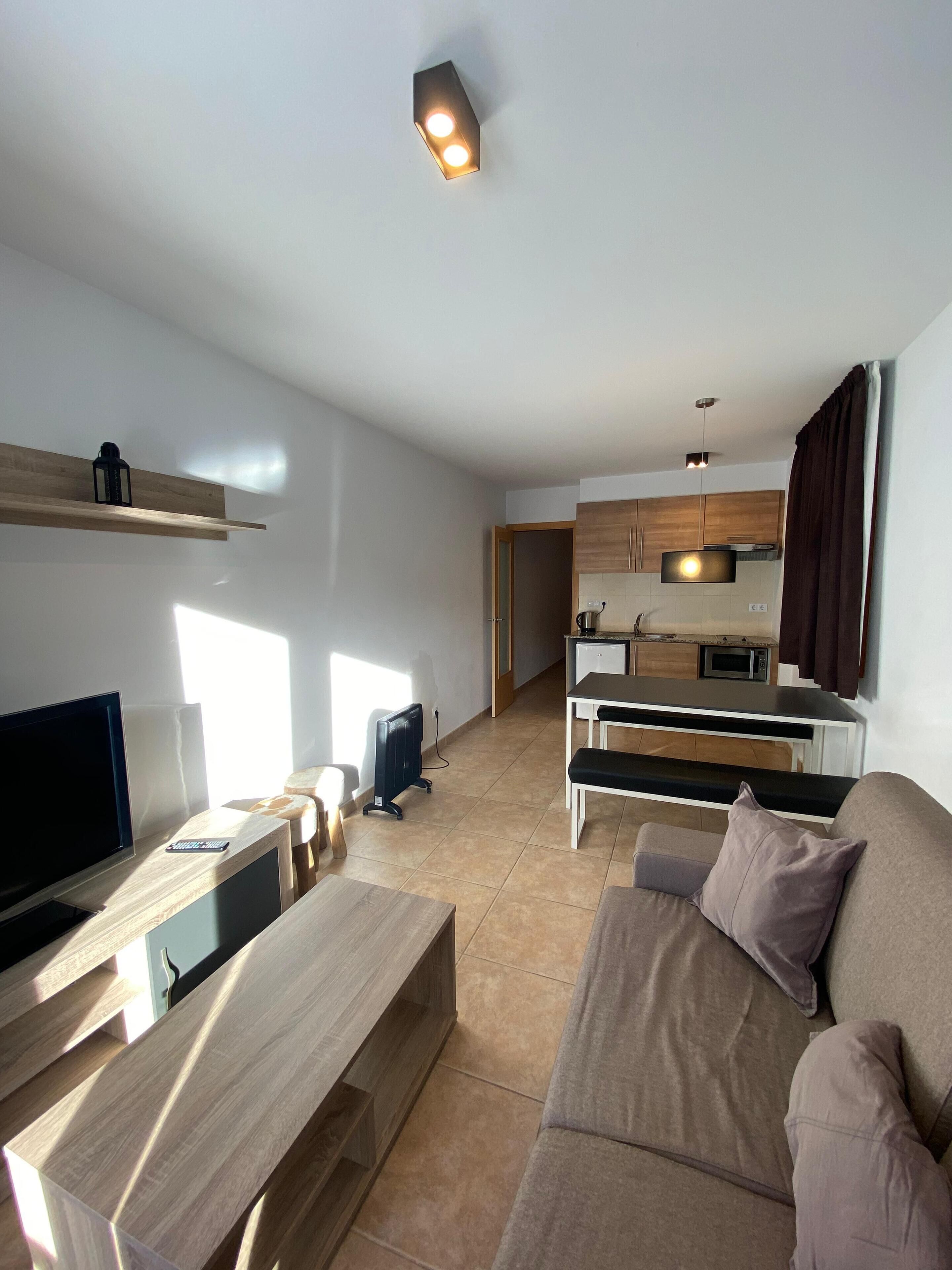 A modern living room with a flat-screen TV, wooden coffee table, and a beige sofa. A kitchen with wooden cabinets and a microwave is visible in the background.