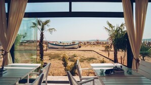 A beach view from a room with a large window, featuring a boat on the sand and palm trees. - Apolonia Resort (Sozopol)