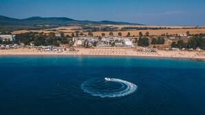 An aerial view of a coastal area with a boat creating a wake in the water, a beach with sun loungers, and a building complex in the background. - Apolonia Resort (Sozopol)