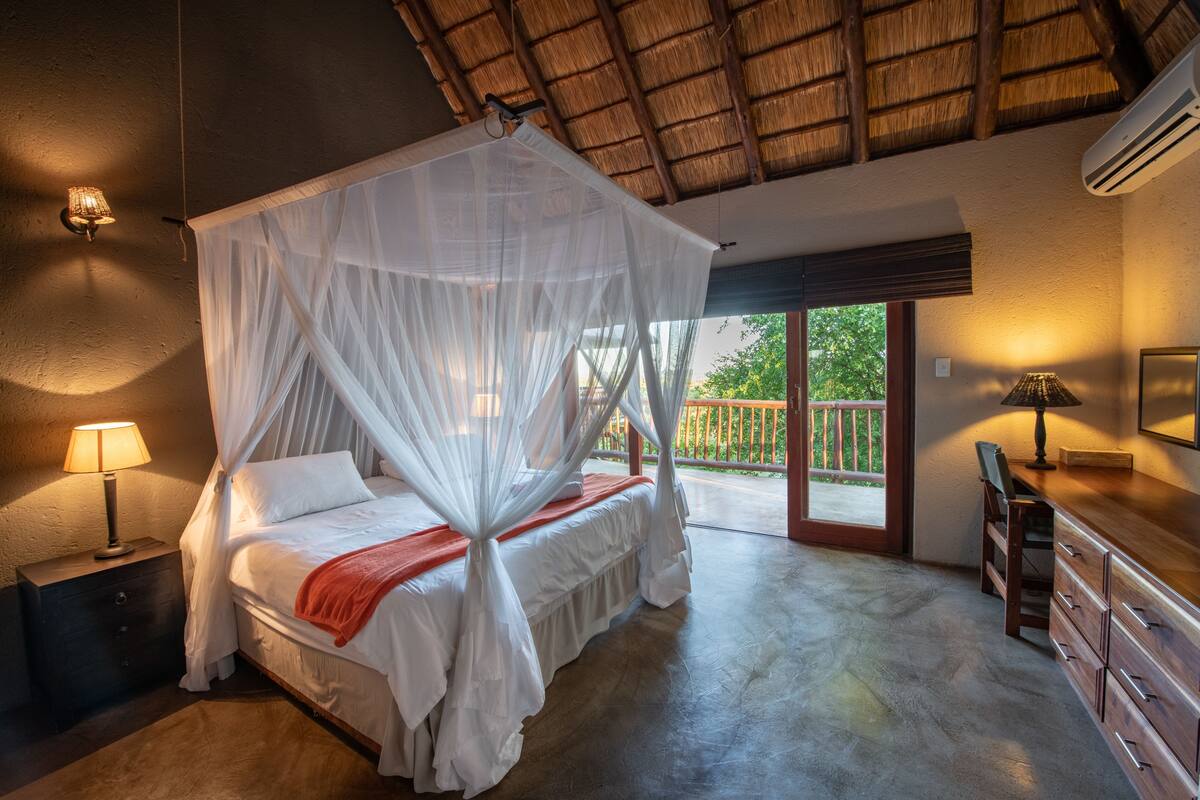 A bedroom with a canopy bed, a nightstand with a lamp, a dresser, and a view of the outdoors.