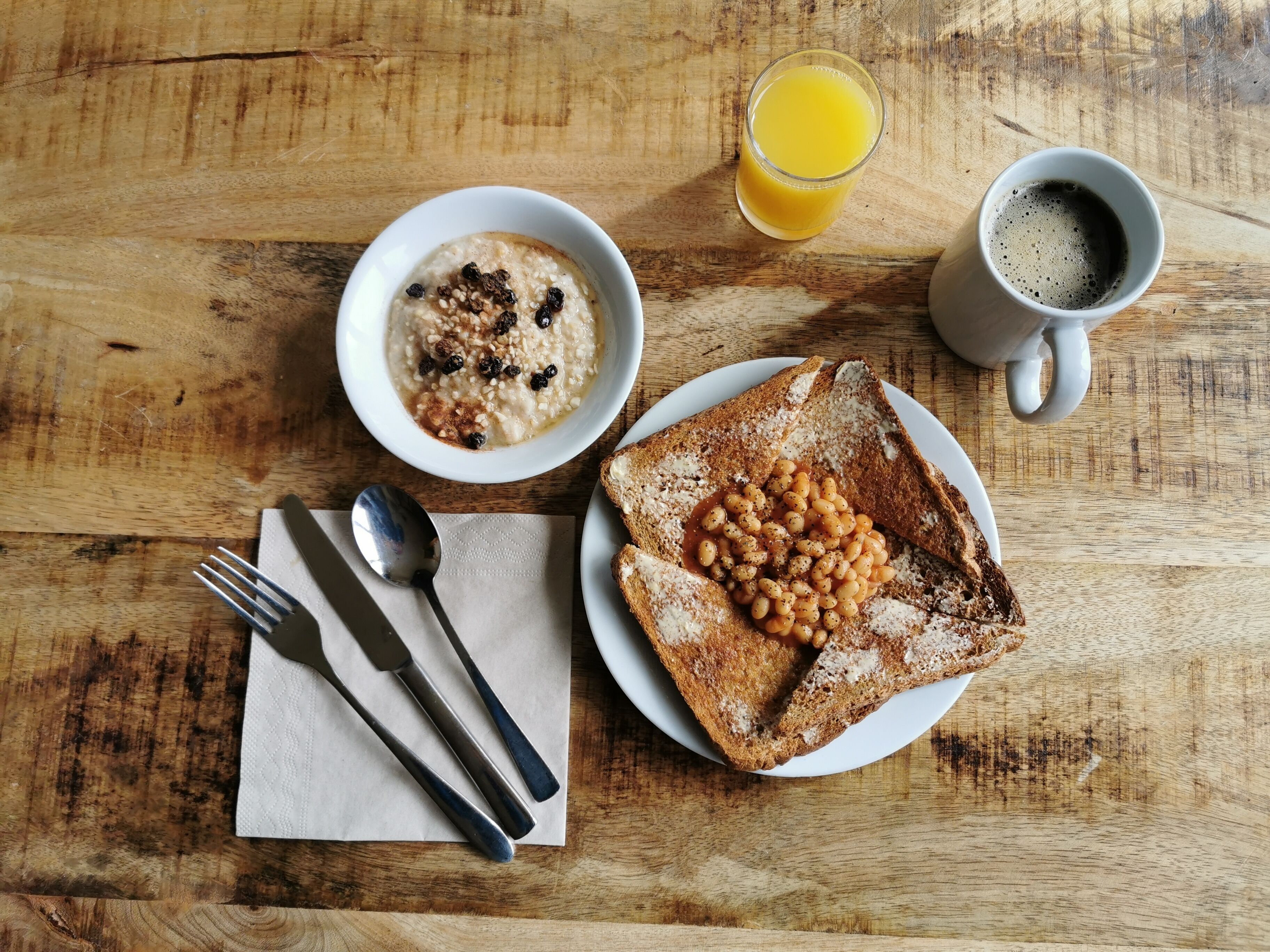 A wooden table with a bowl of oatmeal, toast with baked beans, a glass of orange juice, and a cup of coffee.
